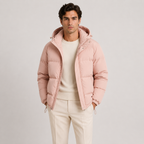 A man wearing a light pink hooded puffer jacket over a cream sweater and cream trousers, standing against a plain light gray studio background.