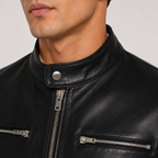 Close-up of a black leather motorcycle jacket worn by a model, showing the stand-up collar with a silver snap button, a front zipper, and zippered chest pockets.