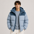 A man wearing a light blue and navy puffer jacket with a corduroy hood, layered over a light blue ribbed sweater and cream pants, against a neutral light gray background.
