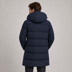 A man viewed from behind wearing a dark navy blue hooded puffer jacket against a plain light gray studio background.