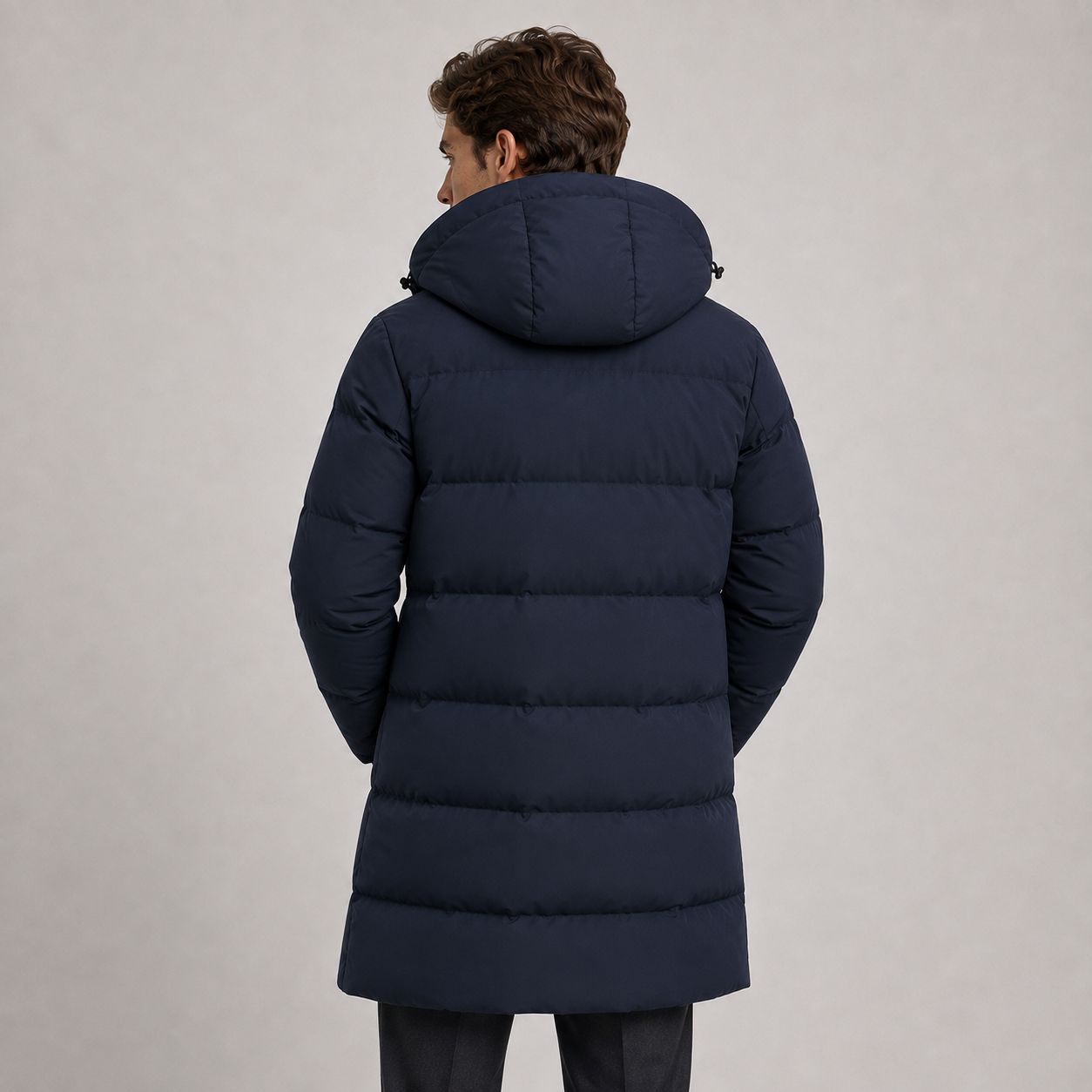 A man viewed from behind wearing a dark navy blue hooded puffer jacket against a plain light gray studio background.