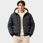 A man wearing a black quilted hooded jacket with a cream sherpa-lined hood, layered over a cream knit sweater and beige pants, against a light gray studio background.
