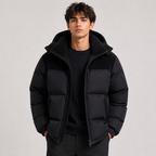 A man wearing a black hooded puffer jacket with black contrast panels on the shoulders and chest, layered over a black ribbed sweater, photographed against a plain light gray studio background.