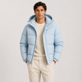 A man wearing a light blue hooded puffer jacket over a cream sweater and cream trousers, standing against a plain light gray studio background.