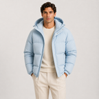 A man wearing a light blue hooded puffer jacket over a cream sweater and cream trousers, standing against a plain light gray studio background.