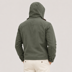 A man viewed from behind wearing an olive green hooded jacket and off-white trousers, standing against a plain light gray studio background.