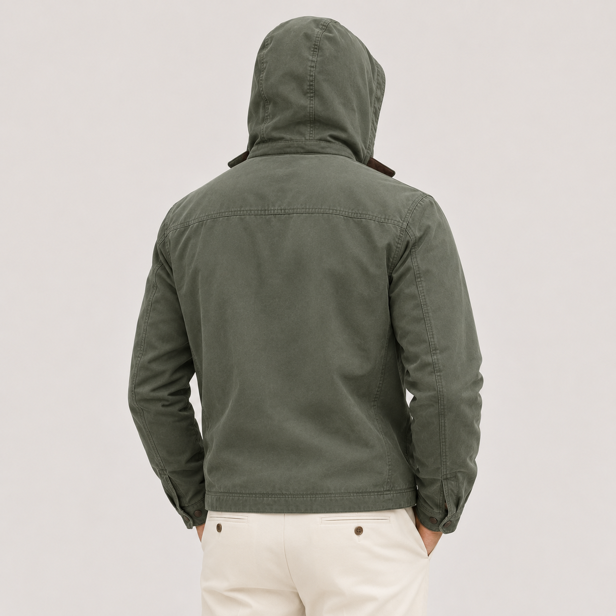 A man viewed from behind wearing an olive green hooded jacket and off-white trousers, standing against a plain light gray studio background.