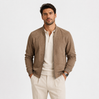 A man wearing a taupe brown suede bomber jacket over a cream polo shirt and cream trousers, standing against a light gray studio background.