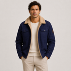 A man wearing a navy blue jacket with a beige sherpa collar, layered over a cream ribbed sweater and beige trousers, standing against a light gray studio background.