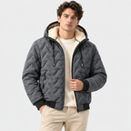 A man wearing a dark gray quilted hooded puffer jacket with a cream sherpa lining, styled over a cream knit sweater and beige trousers, against a light gray studio background.