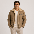 A man wearing a tan corduroy-trimmed jacket over a cream ribbed sweater and beige trousers, standing against a light gray studio background.