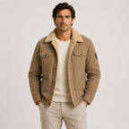 A man wearing a tan corduroy-trimmed jacket over a cream ribbed sweater and beige trousers, standing against a light gray studio background.