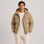 A man wearing a light brown hooded puffer jacket over a cream crewneck sweater and cream trousers, standing against a light gray studio background.