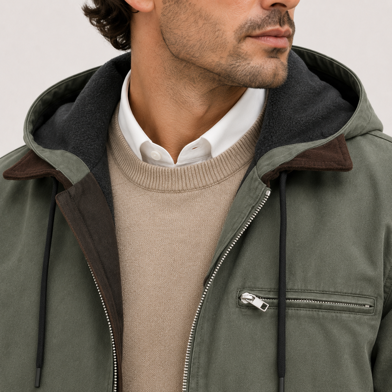 Close-up of a man wearing an olive green hooded jacket with a dark gray fleece-lined hood and brown collar trim, layered over a beige sweater and white collared shirt.