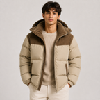 A man wearing a beige and brown puffer jacket with a corduroy collar and hood, layered over a beige ribbed sweater and cream pants, photographed against a light gray studio background.