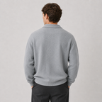 A man viewed from behind wearing a light gray knit sweater and dark trousers, standing against a plain light gray studio background.