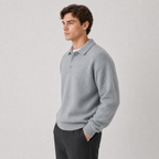 A man wearing a light gray knit polo sweater and dark trousers, standing against a plain light gray studio background.