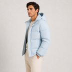 A man wearing a light blue hooded puffer jacket over a cream sweater and beige trousers, standing against a plain light gray studio background.
