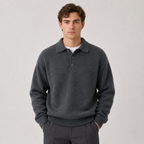 A man wearing a dark gray knit polo sweater over a white undershirt, paired with dark gray trousers, standing against a light gray studio background.