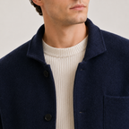 Close-up of a man wearing a navy blue wool-blend jacket over a cream ribbed knit sweater, against a light gray studio background.