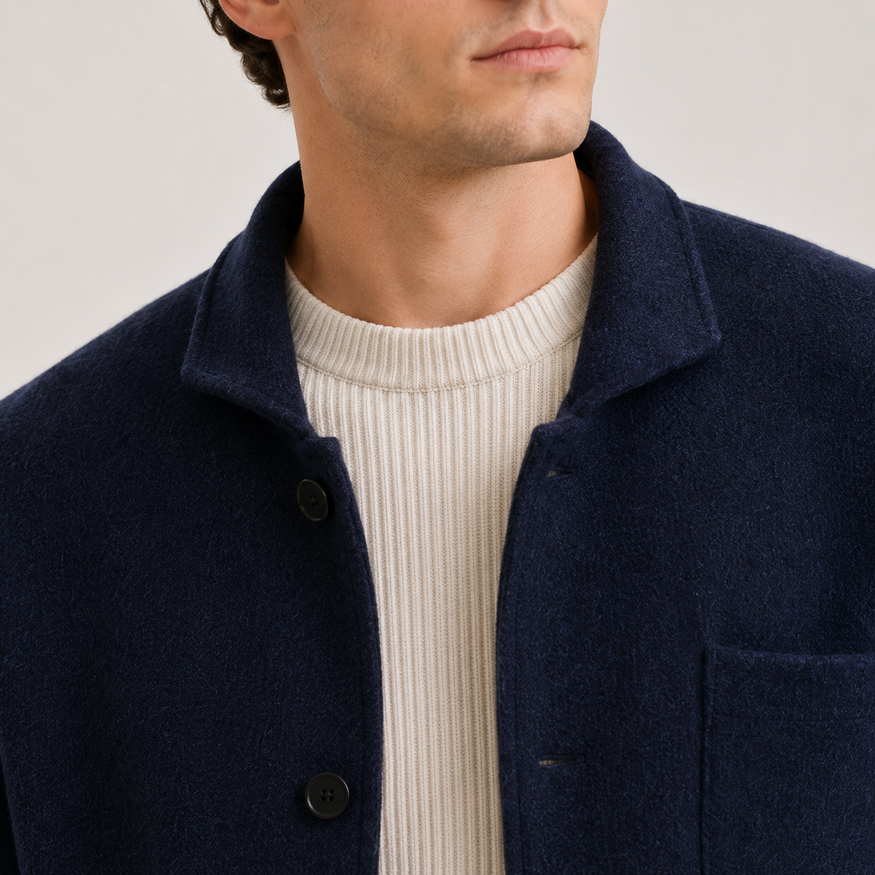 Close-up of a man wearing a navy blue wool-blend jacket over a cream ribbed knit sweater, against a light gray studio background.