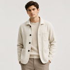 A man wearing a light beige textured cardigan over a cream ribbed sweater and beige trousers, standing against a plain light gray studio background.
