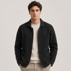 A man wearing a dark charcoal wool-blend button-front cardigan over a cream ribbed sweater and beige trousers, standing against a light gray studio background.