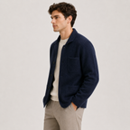 A man wearing a navy blue knit cardigan over a cream crewneck sweater and beige trousers, standing against a light gray studio background.