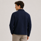 A man shown from the back wearing a dark navy blue knit sweater and light beige trousers, standing against a plain light gray studio background.