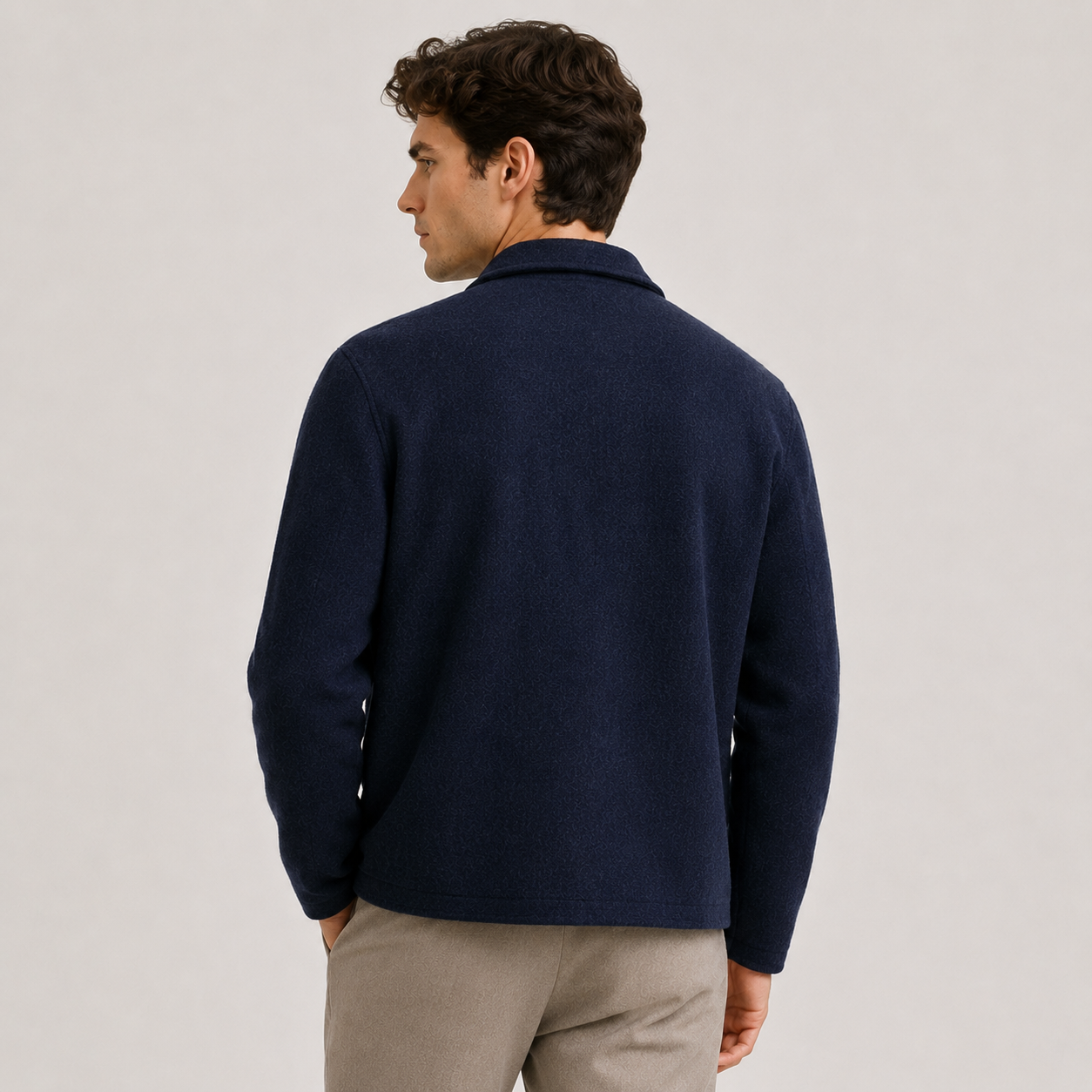 A man shown from the back wearing a dark navy blue knit sweater and light beige trousers, standing against a plain light gray studio background.