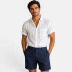 Man wearing a white short-sleeve shirt and navy shorts on a plain background