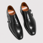 A pair of black leather double monk-style dress shoes with silver-tone buckle straps, shown from a top-down angle on a light gray background.