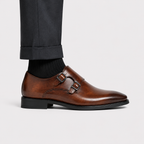 A brown leather double monk strap shoe with black soles, shown on a model's foot with black trousers and a black sock against a light gray background.