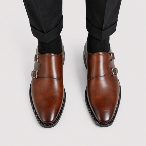 A pair of men's brown leather double monk shoes with square toes and double buckle straps, shown on a model wearing black trousers and black socks against a light gray background.