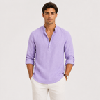 A man wearing a light purple long-sleeve linen henley shirt with white pants, standing against a light gray studio background.