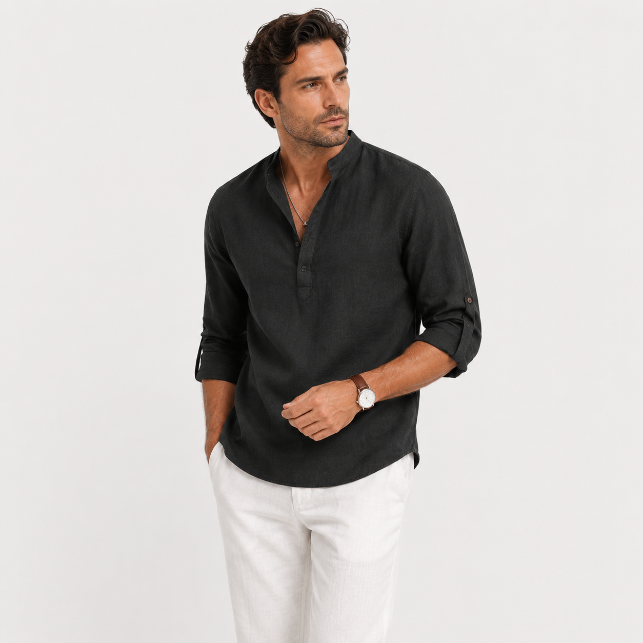 A man wearing a black linen long-sleeve shirt with rolled-up sleeves and white trousers, standing against a light gray studio background.