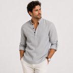 A man wearing a light gray long-sleeve linen henley shirt with rolled-up sleeves and white trousers, standing against a plain light gray background.