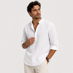 A man wearing a white long-sleeve linen shirt with a split collar and buttoned placket, paired with light beige trousers, photographed against a solid light gray background.