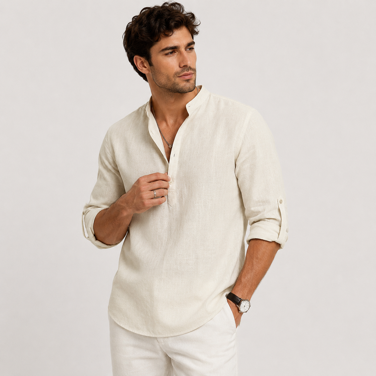 A man wearing a light beige linen long-sleeve shirt with a buttoned collar and white trousers, standing against a light gray studio background.