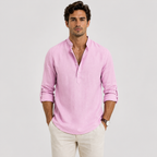 A man wearing a light pink long-sleeve linen henley shirt with rolled-up sleeves and cream trousers, standing against a light gray studio background.