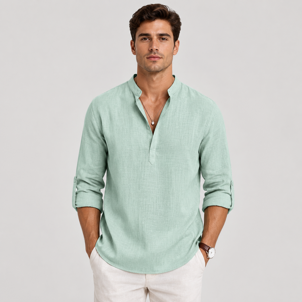 A man wearing a light mint green linen henley shirt with rolled-up sleeves and white pants, standing against a neutral light gray background.