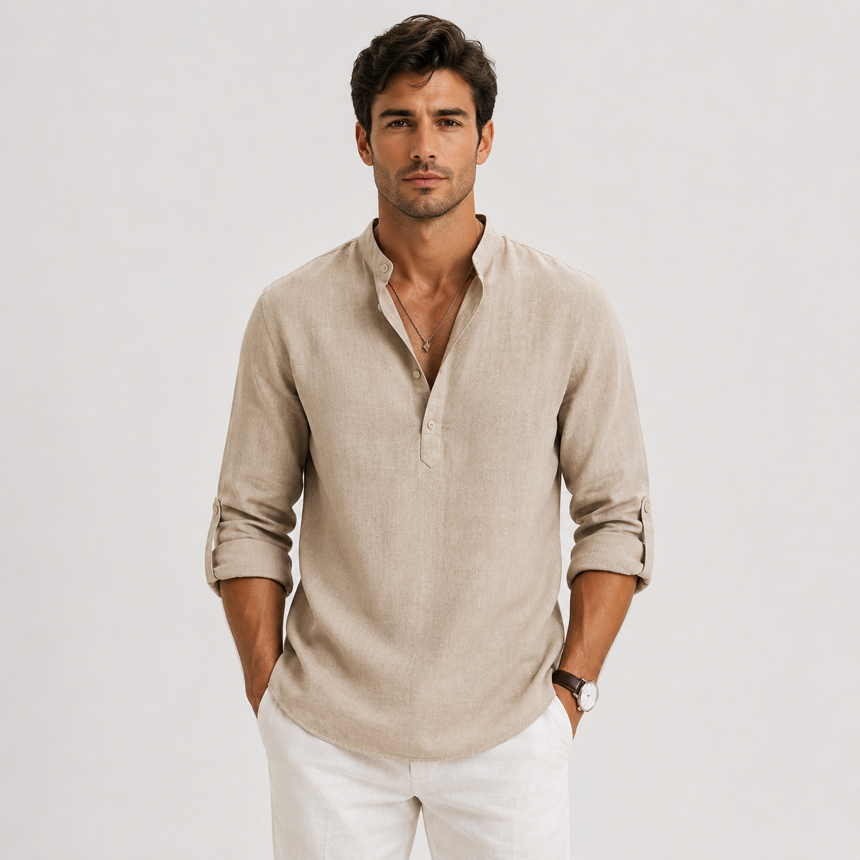 A man wearing a light beige linen henley shirt with rolled-up sleeves and white trousers, standing against a light gray studio background.