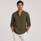 A man wearing an olive green long-sleeve linen henley shirt with white trousers, standing against a light gray studio background.