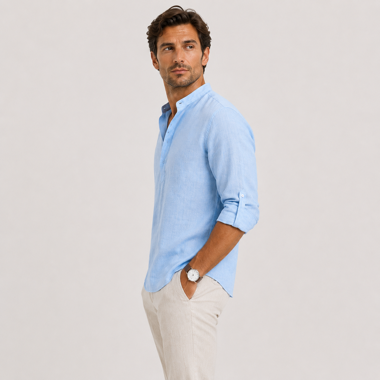 A man wearing a light blue long-sleeve linen shirt with rolled-up sleeves and beige trousers, standing against a light gray studio background.