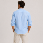 A man viewed from the back wearing a light blue long-sleeve button-down shirt with rolled-up sleeves and beige trousers, set against a plain light gray studio background.
