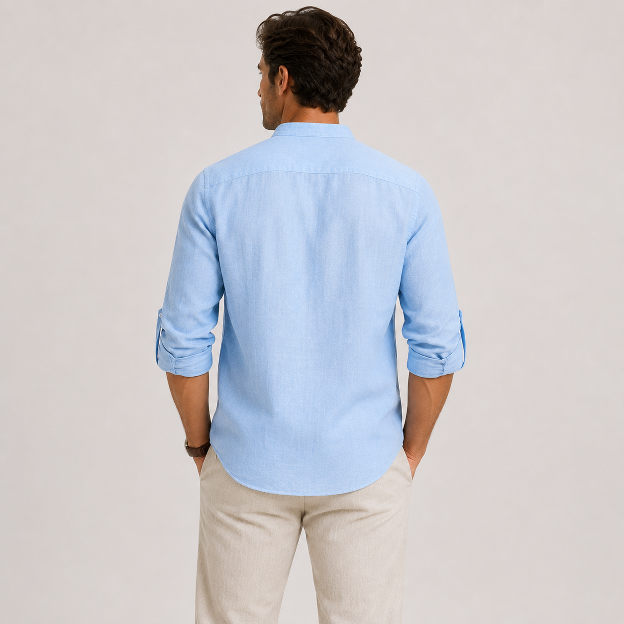 A man viewed from the back wearing a light blue long-sleeve button-down shirt with rolled-up sleeves and beige trousers, set against a plain light gray studio background.