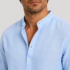 A close-up of a man wearing a light blue linen button-down shirt with a mandarin collar, shown against a neutral light gray background.
