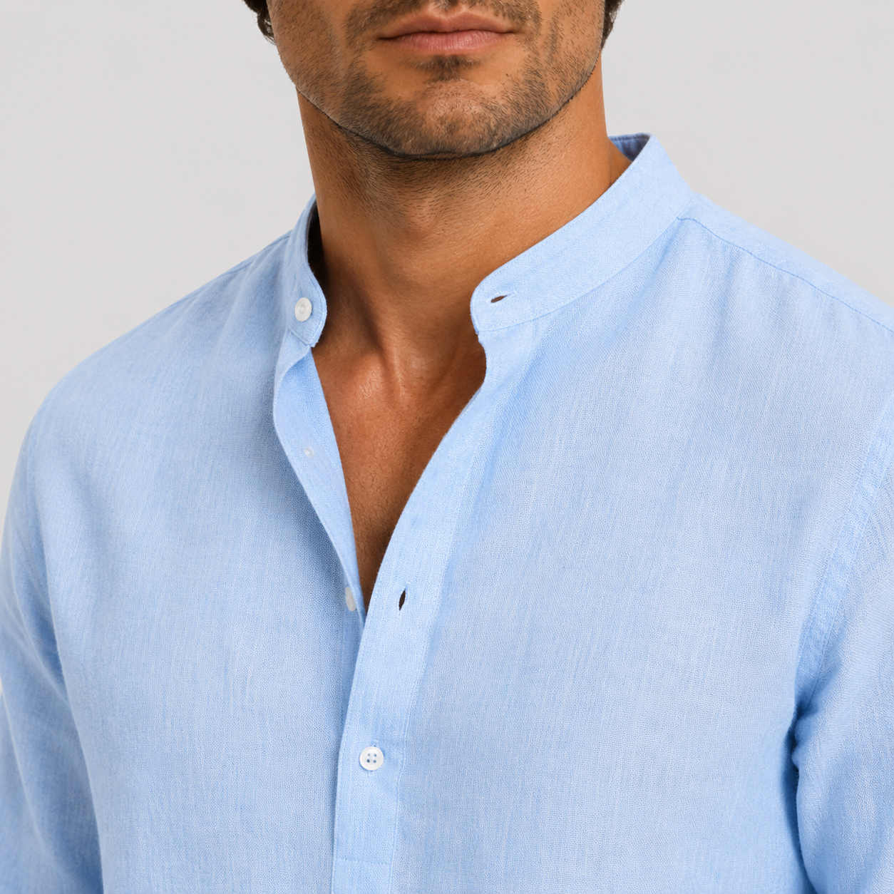 A close-up of a man wearing a light blue linen button-down shirt with a mandarin collar, shown against a neutral light gray background.