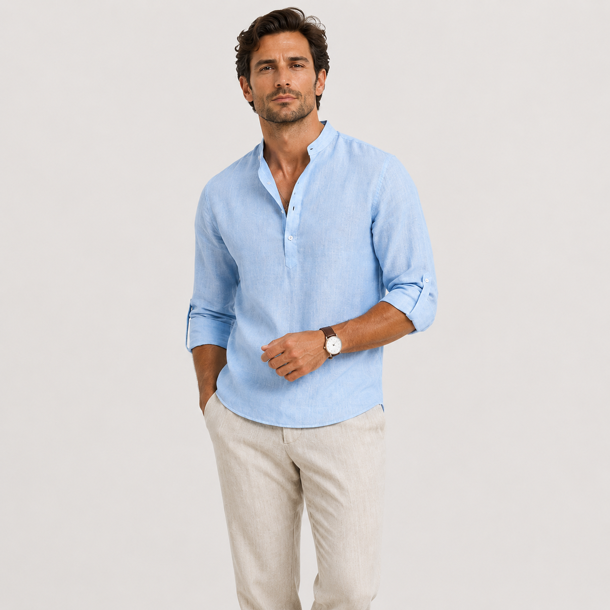 A man modeling a light blue linen button-down shirt with rolled-up sleeves and beige trousers, standing against a plain light gray studio background.