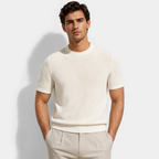 A man wearing a short-sleeve textured knit crewneck sweater in off-white, paired with light beige trousers, standing against a light gray studio background.
